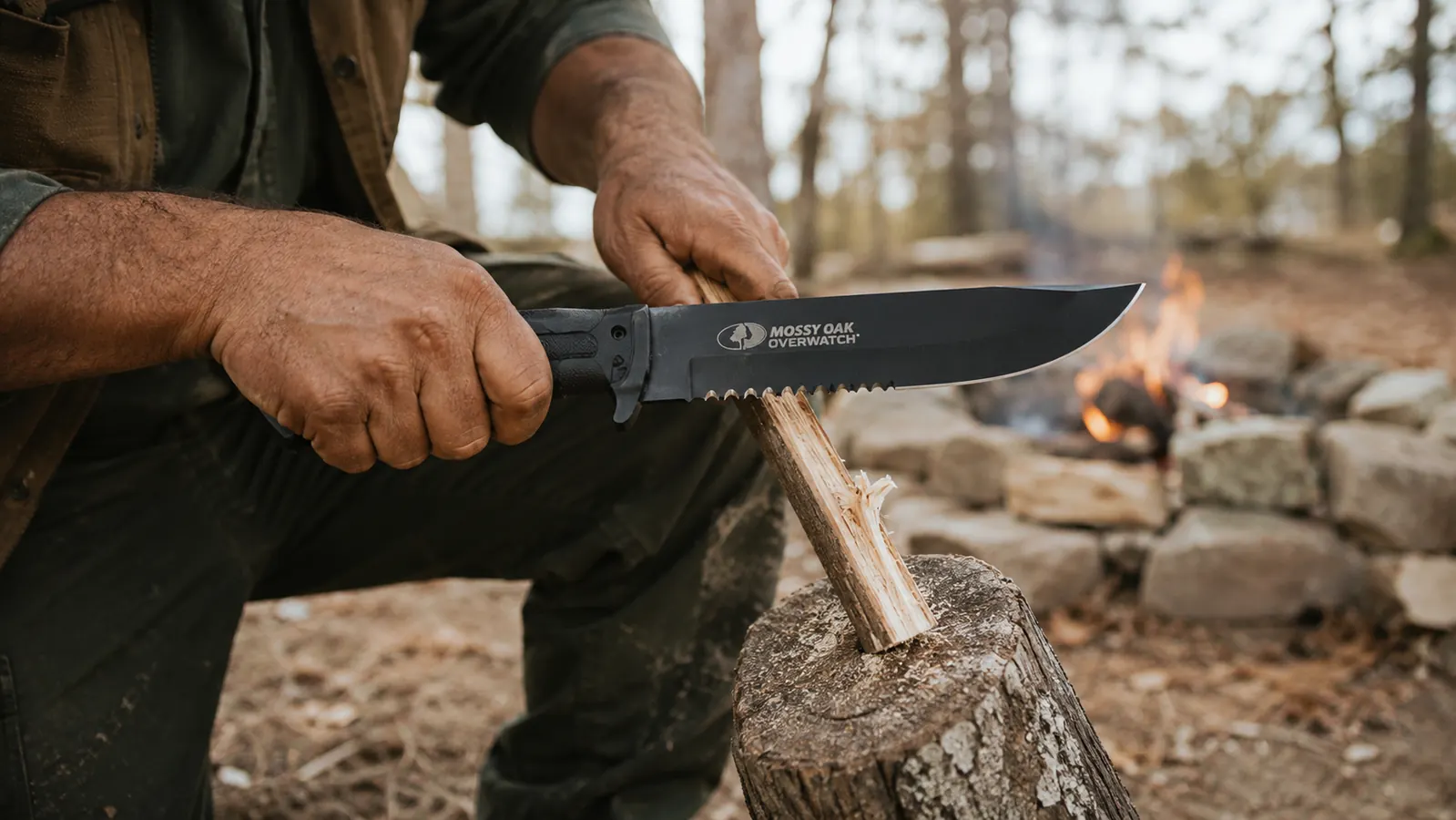 A hand gripping the Mossy Oak fixed blade knife while batoning a small piece of firewood at a camp fire ring