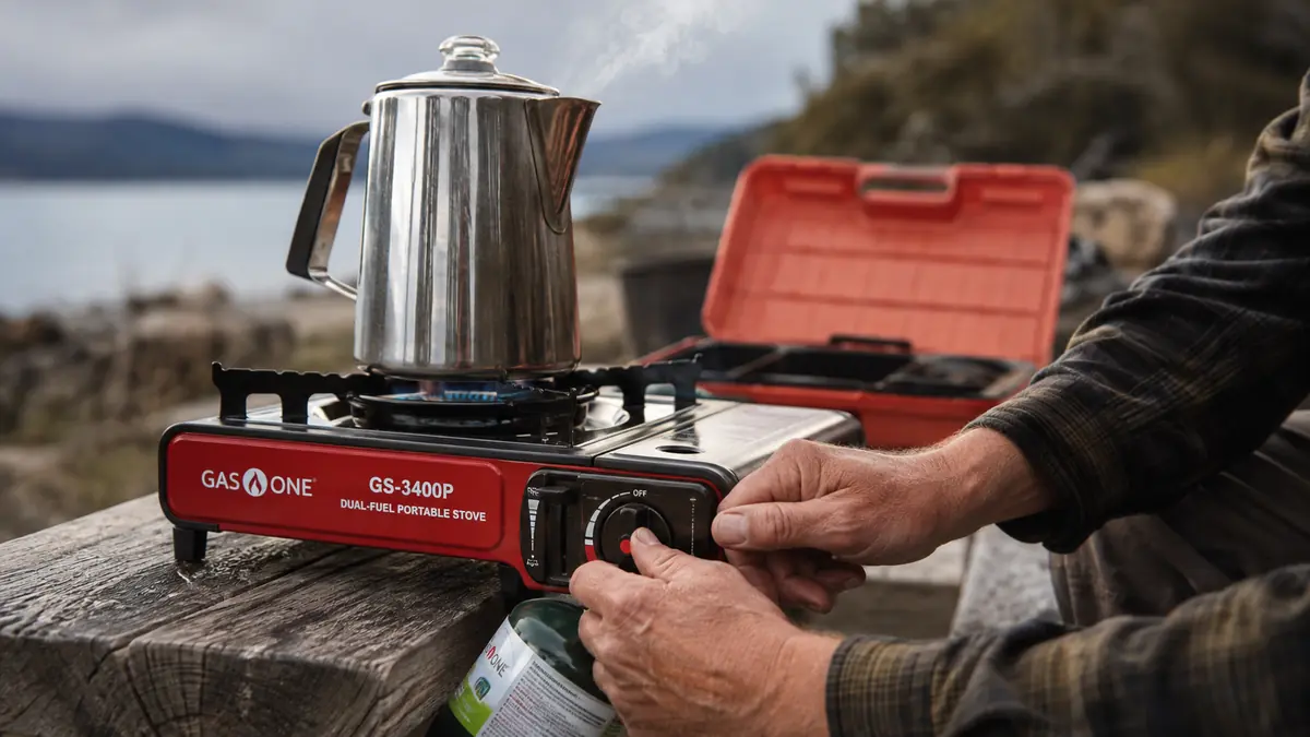 Hands lighting the Gas One GS-3400P portable stove with a percolator coffee pot set on the burner