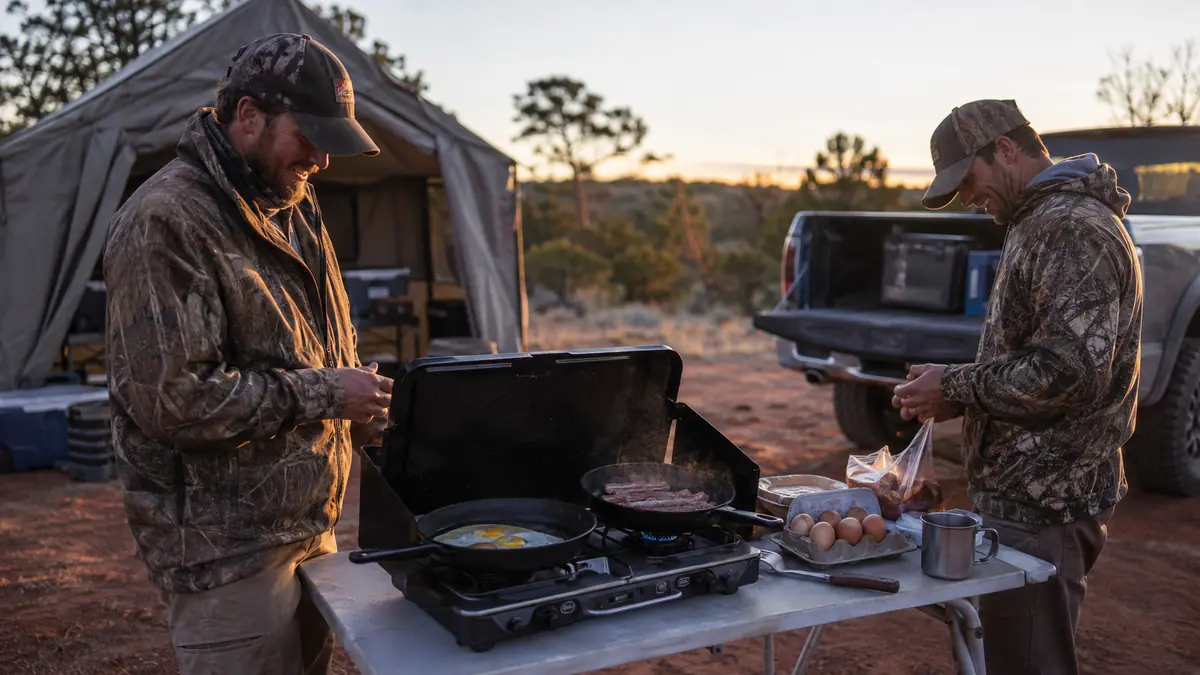 Two hunters cooking a full breakfast spread on a two-burner propane stove at a base camp