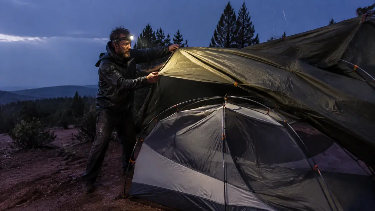 Camper pitching a dome tent rainfly in steady rain at a forest campsite